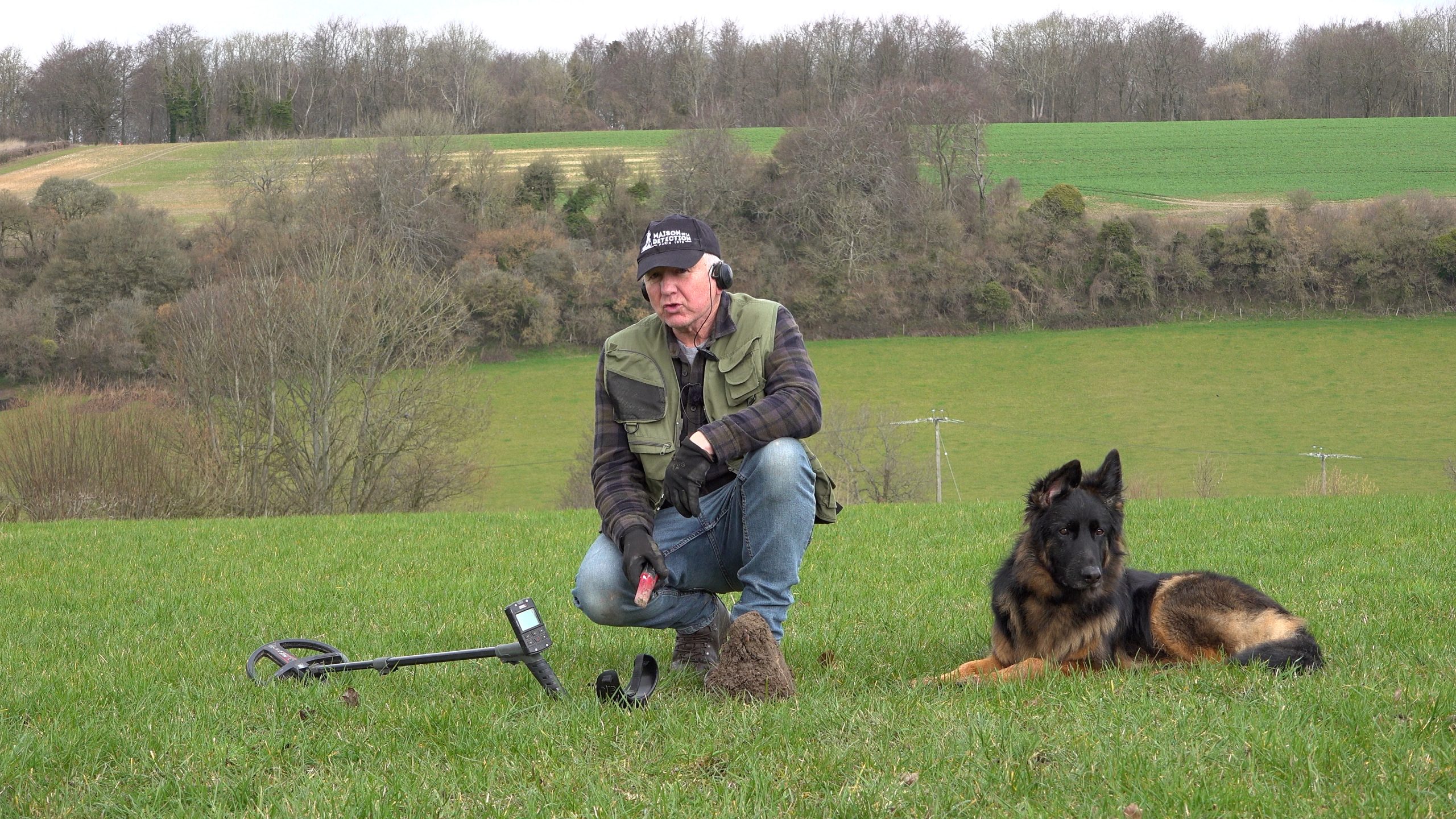 Man in a field with a metal detector next to a large German Shepherd with hills in the background